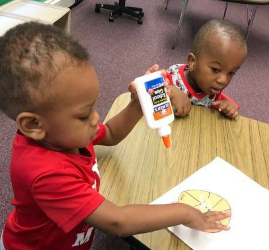 Child holding a bottle of glue over paper, another child watching