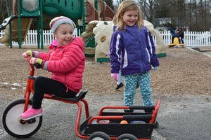Child on a tricycle with a wagon attached, another child standing nearby
