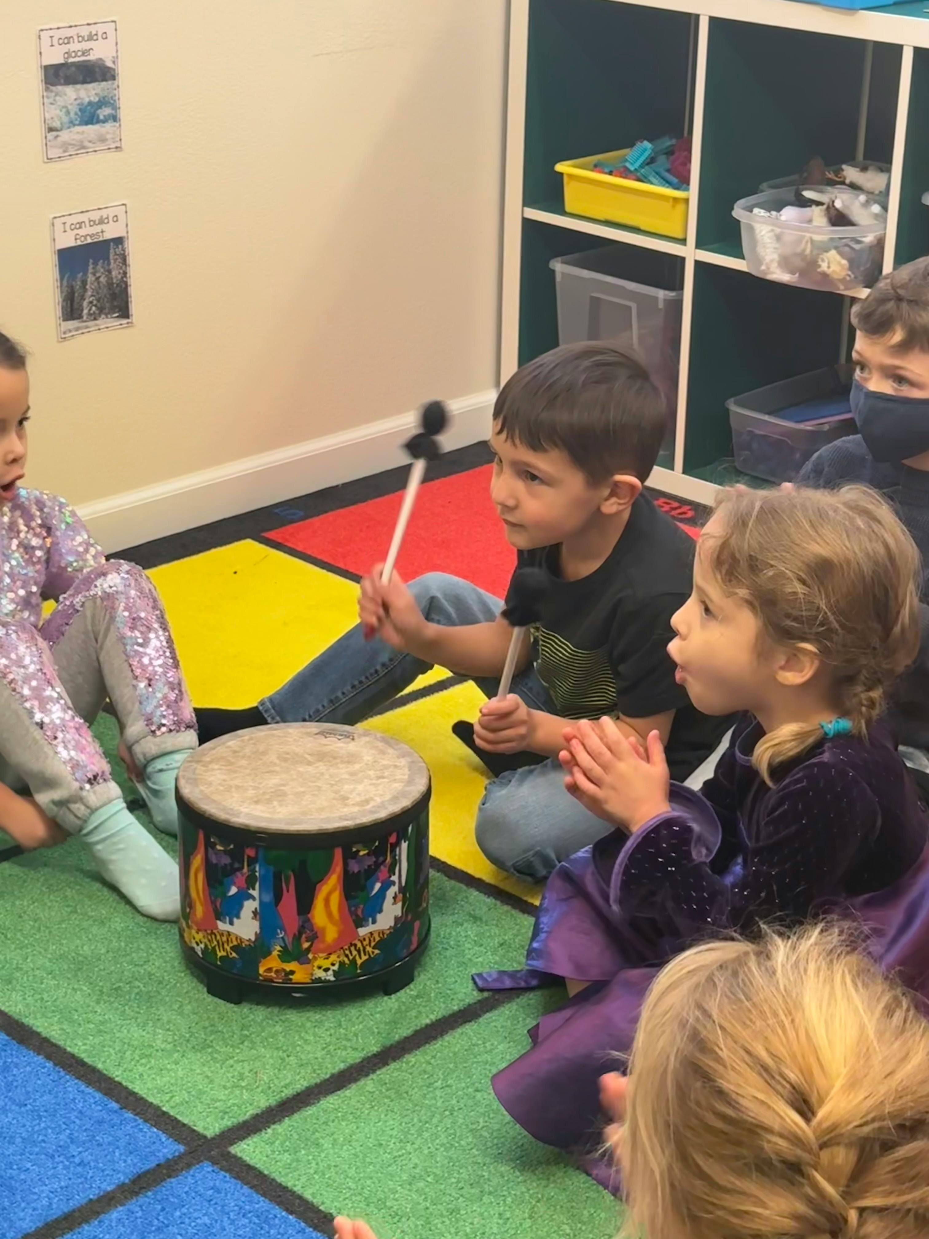 Child playing a drum with a mallet, other children watching