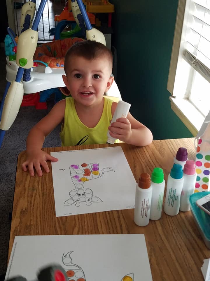 Child holding a white object while sitting at a table with coloring pages and paint bottles