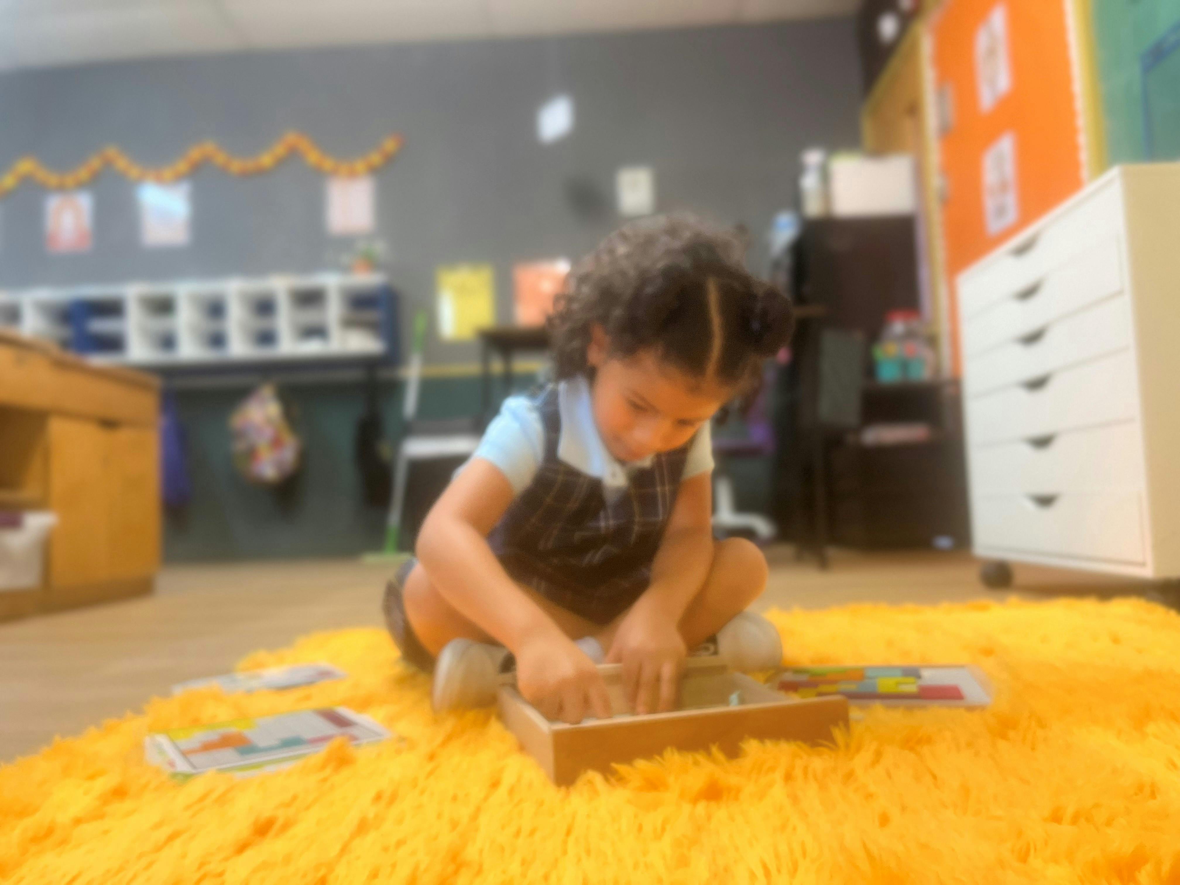 Child playing with a wooden tray on a yellow rug in a classroom