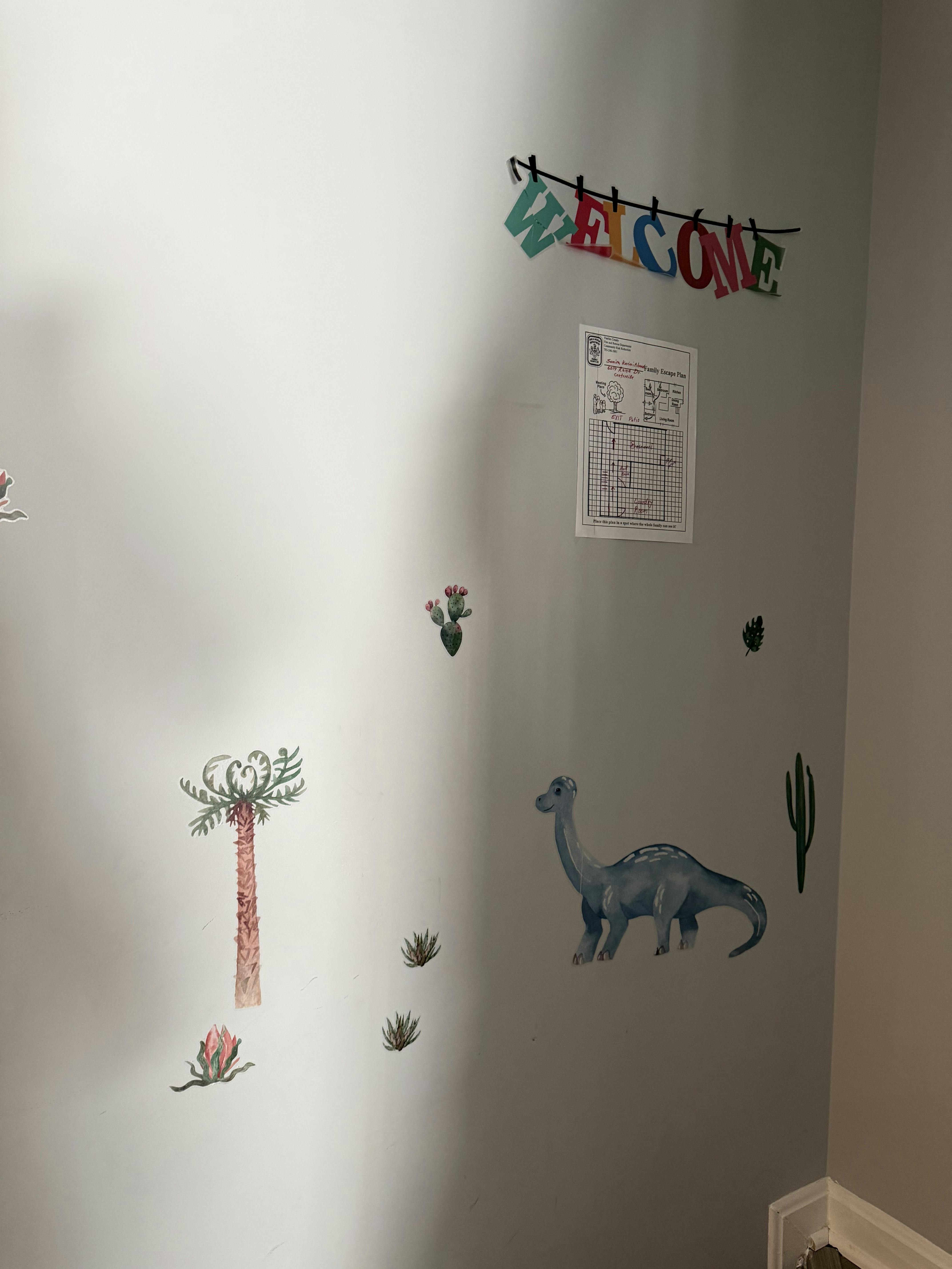 White wall with 'WELCOME' banner, 'Family Escape Plan' chart, dinosaur, and plants