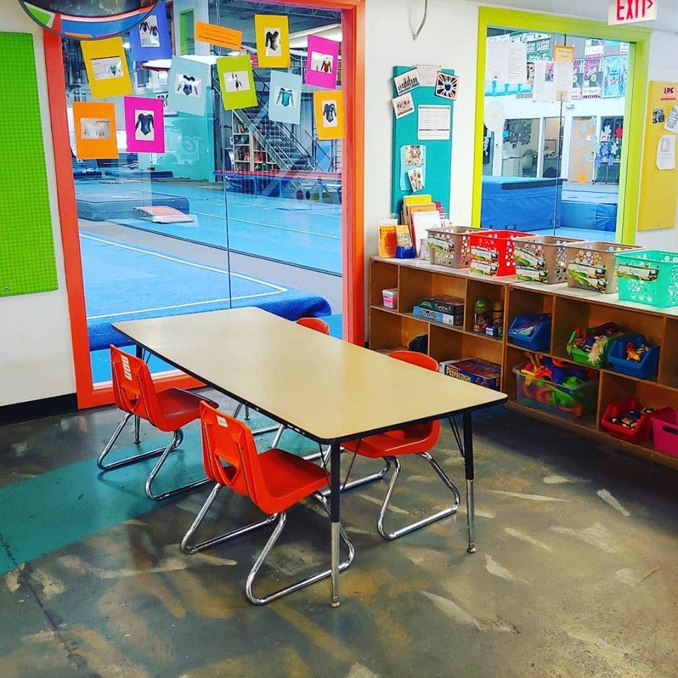 Classroom with a table and chairs, shelves with toys, and a view into a gym