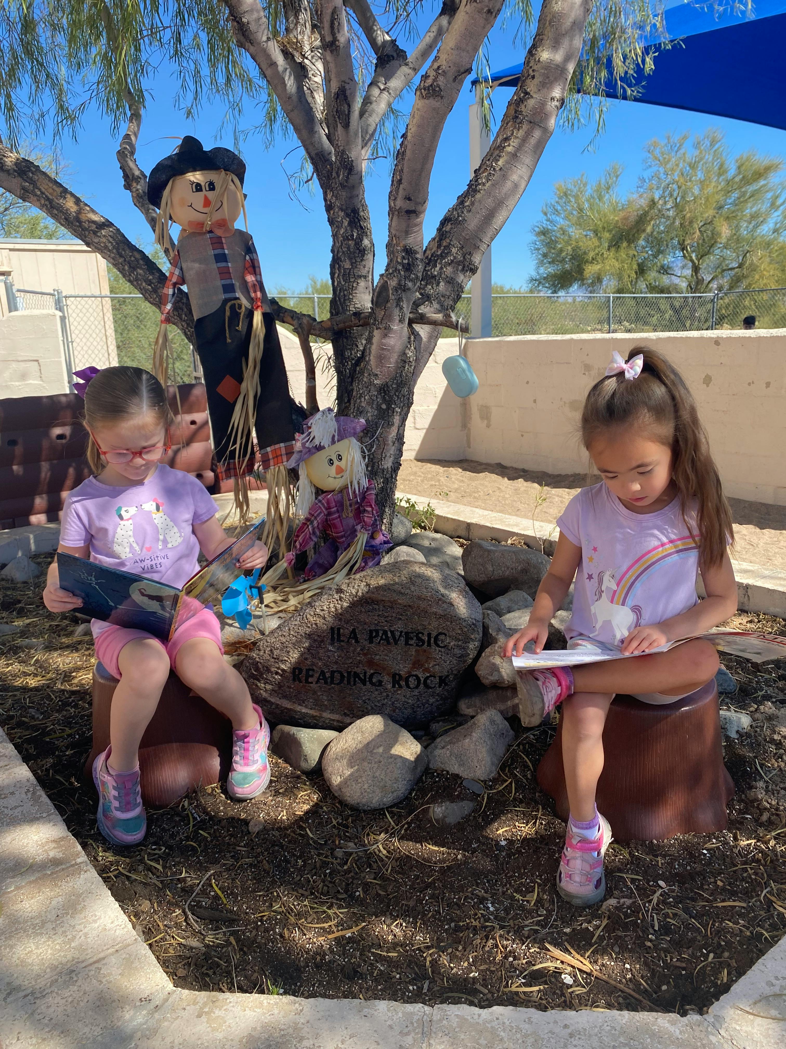 Two children sitting outdoors reading books next to scarecrow decorations