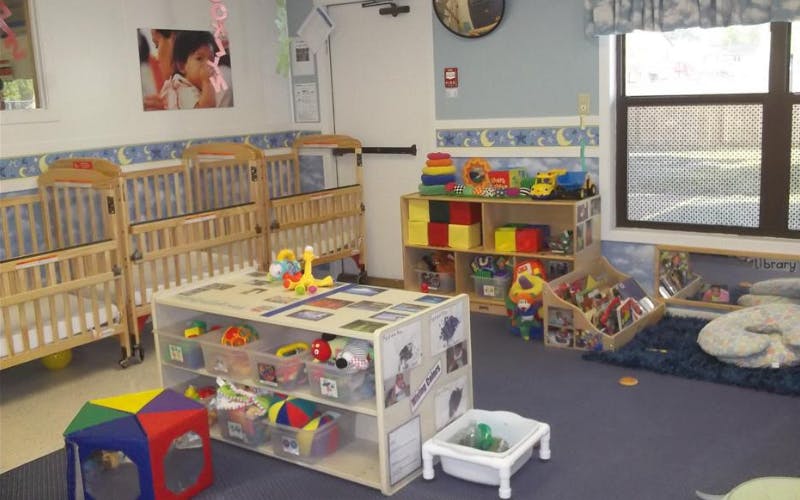 A brightly lit nursery with several cribs, shelves full of toys, and a window