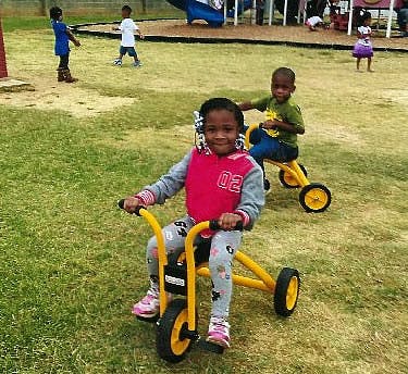 Two children riding tricycles on grass, other children in background