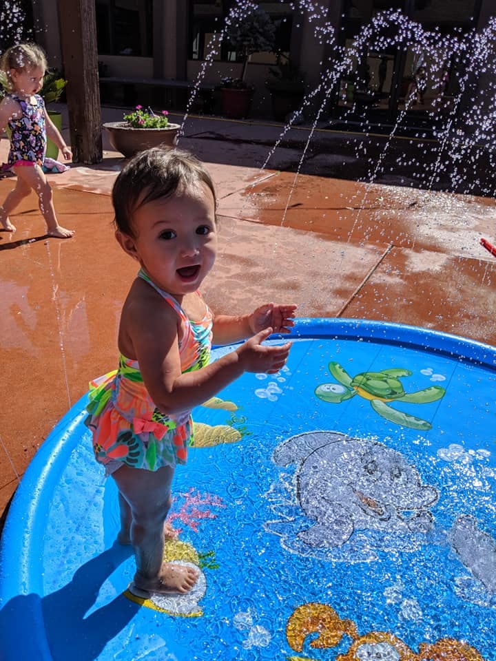 Child in a swimsuit standing in a shallow blue pool with water spraying