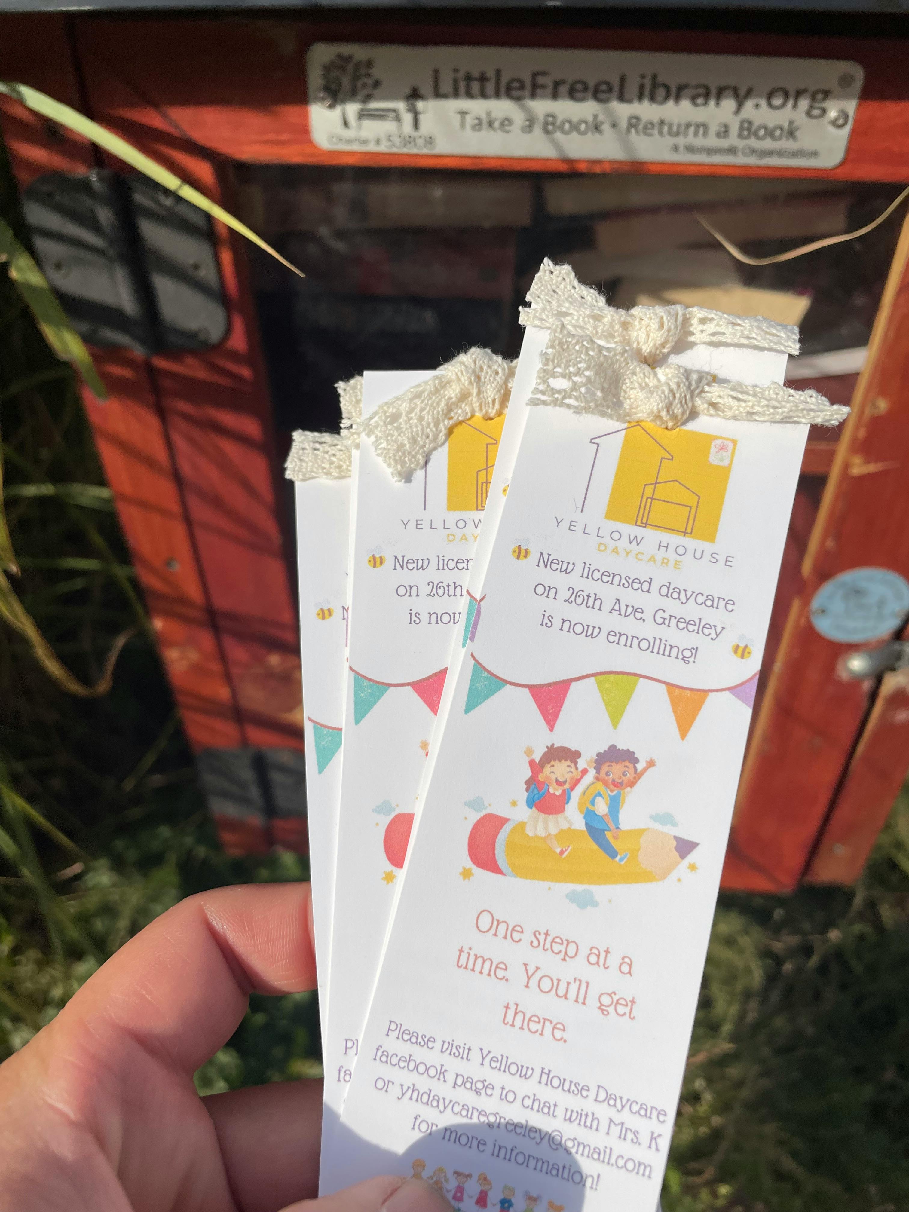 Hand holding bookmarks in front of a Little Free Library