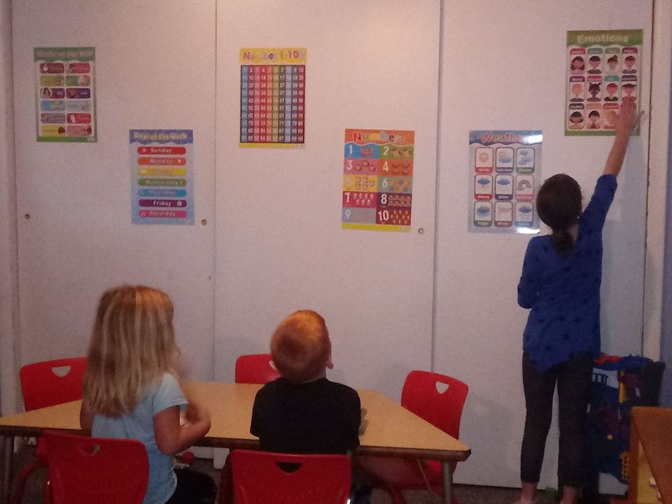 Three children in a classroom, one reaching up to a poster on the wall