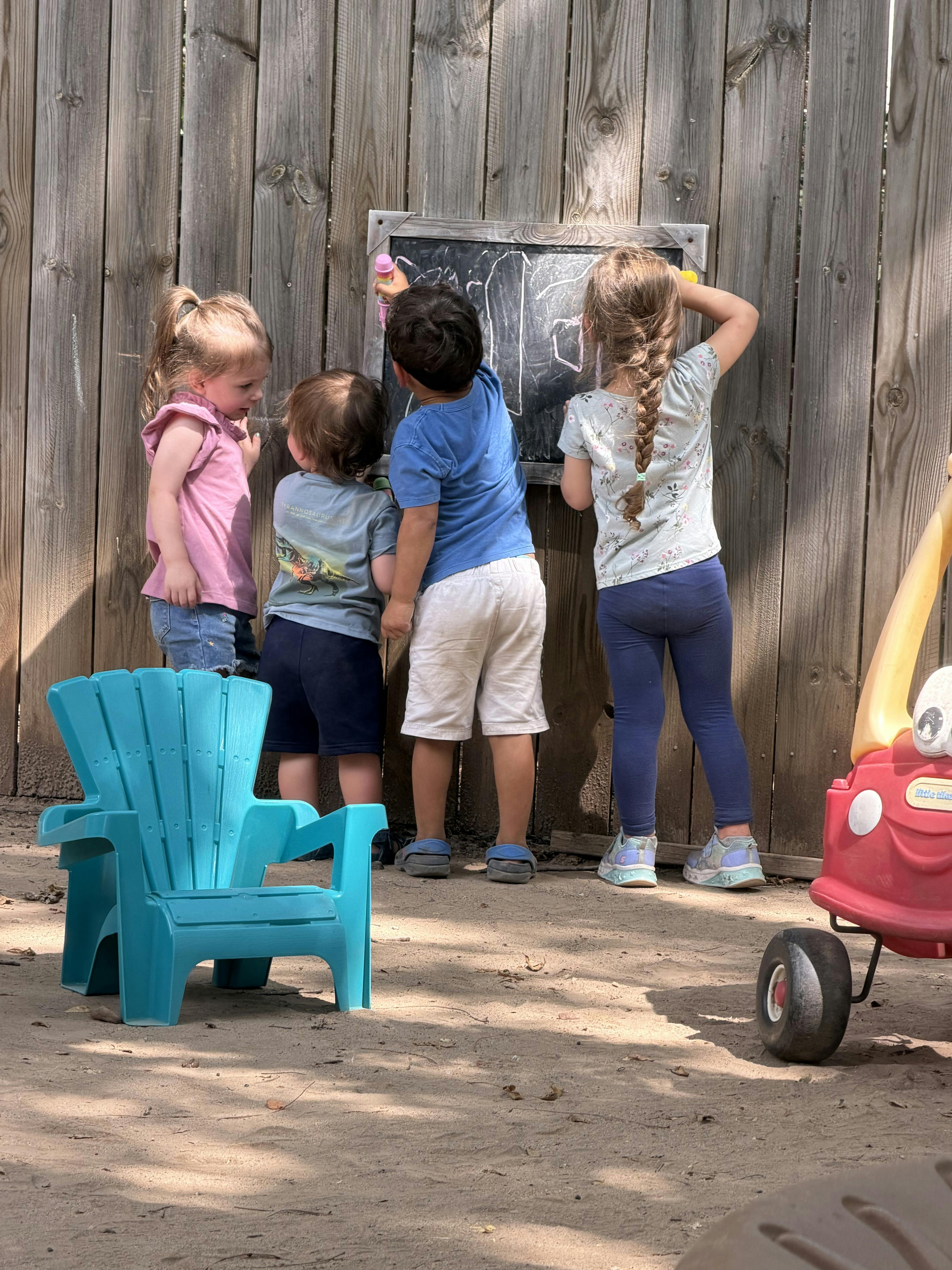 Four children drawing on a chalkboard attached to a wooden fence