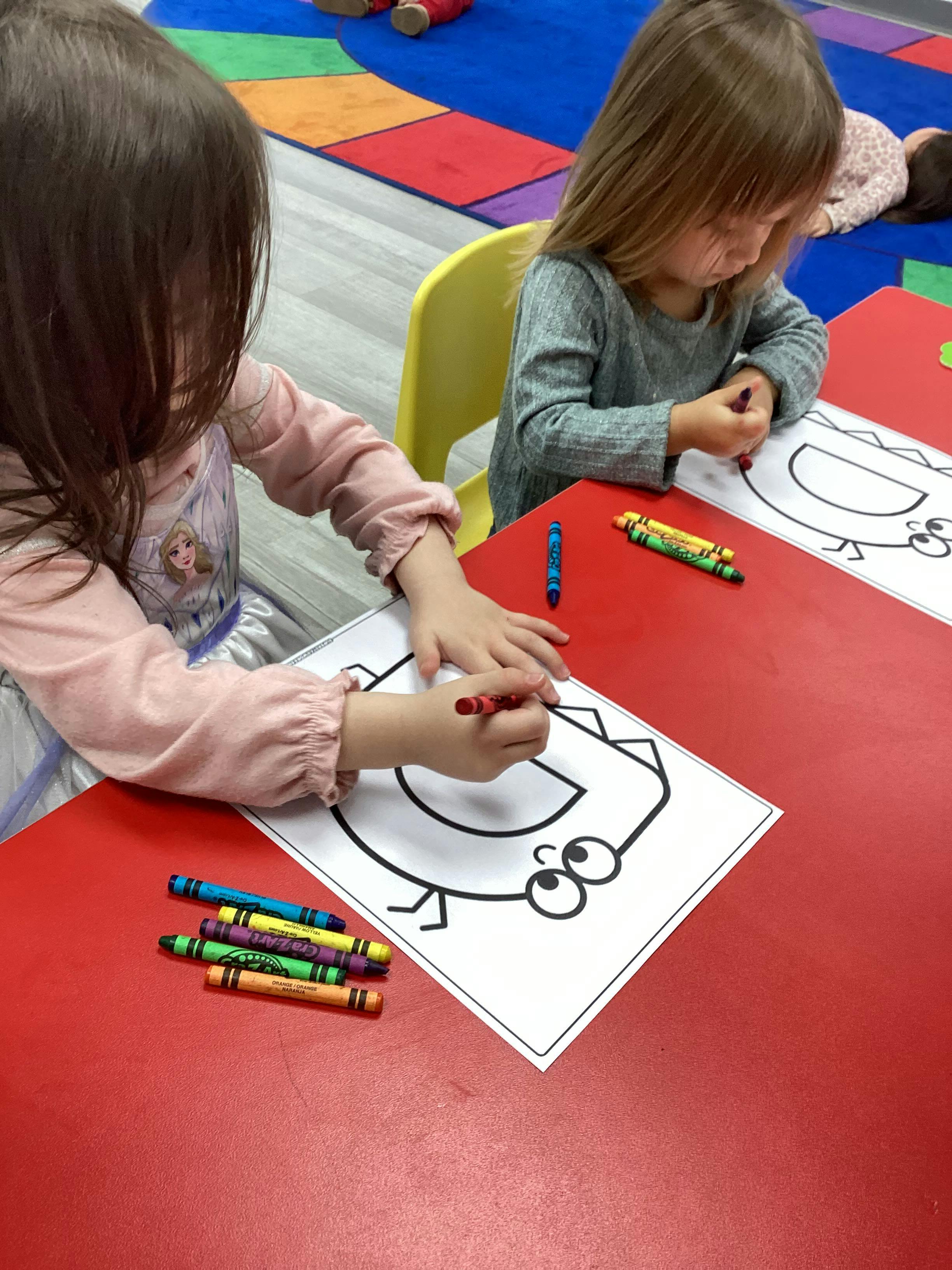 Two children coloring a letter 'D' dinosaur on a red table