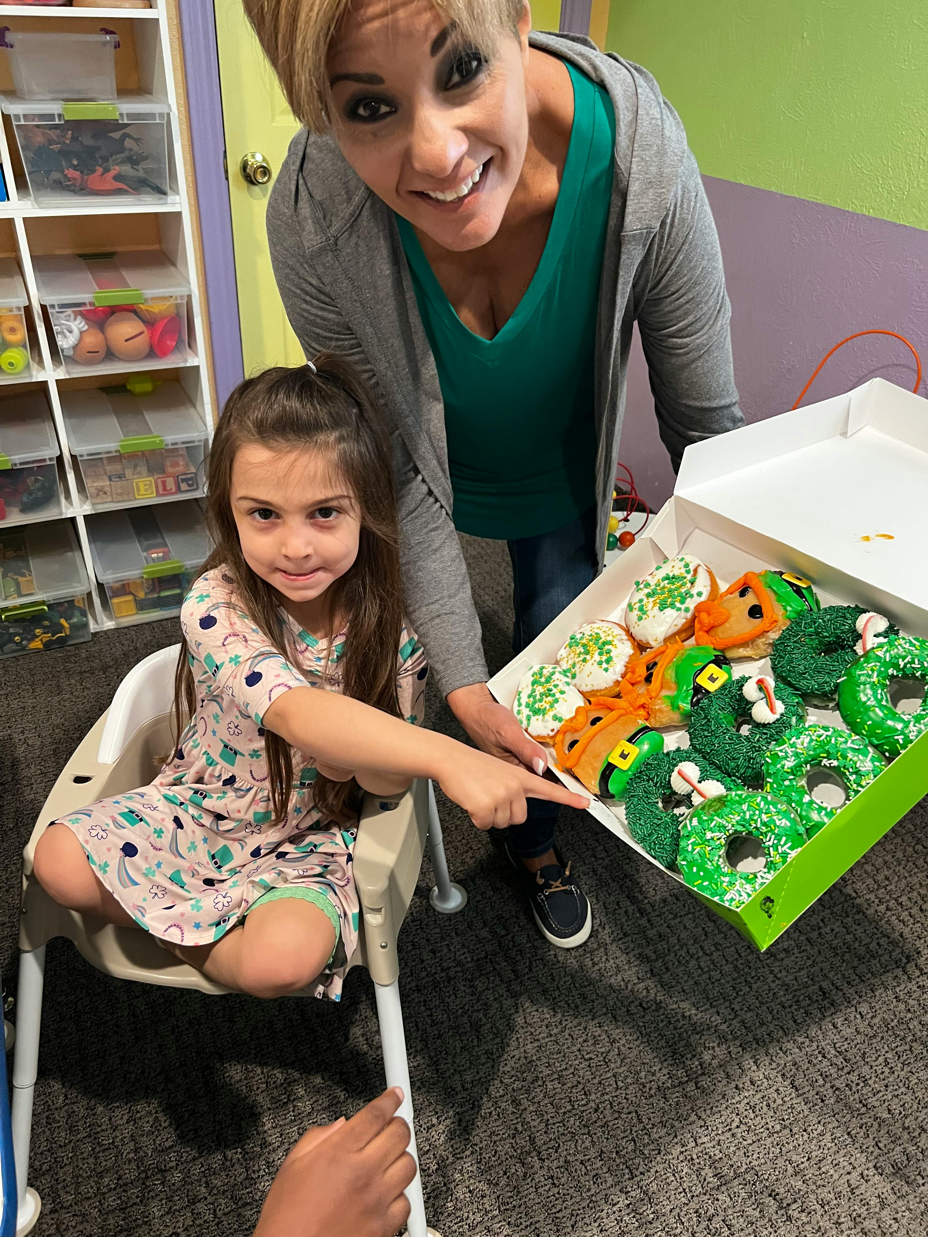 Person holding a box of decorated donuts, child pointing at donuts
