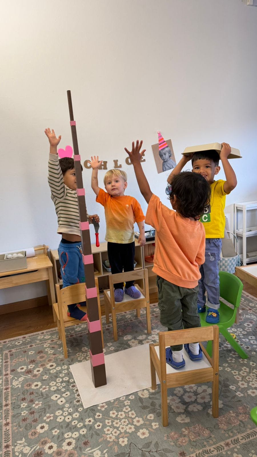Children standing on chairs with hands raised next to a tall block tower