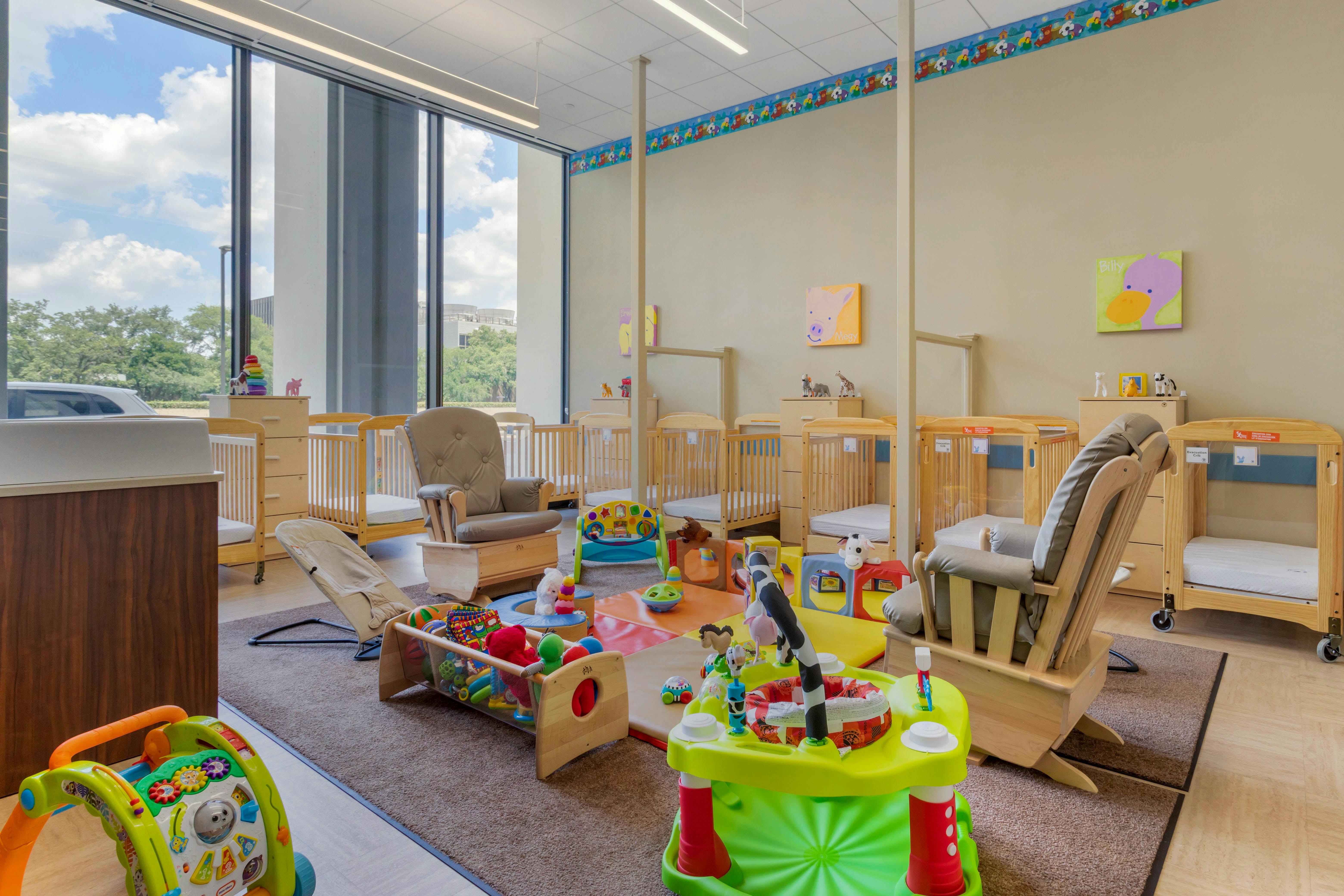 A brightly lit nursery room with multiple cribs, rockers, and infant play items