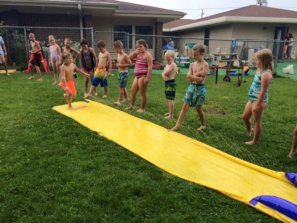 Children in swimsuits standing around a yellow slip and slide on a grassy lawn