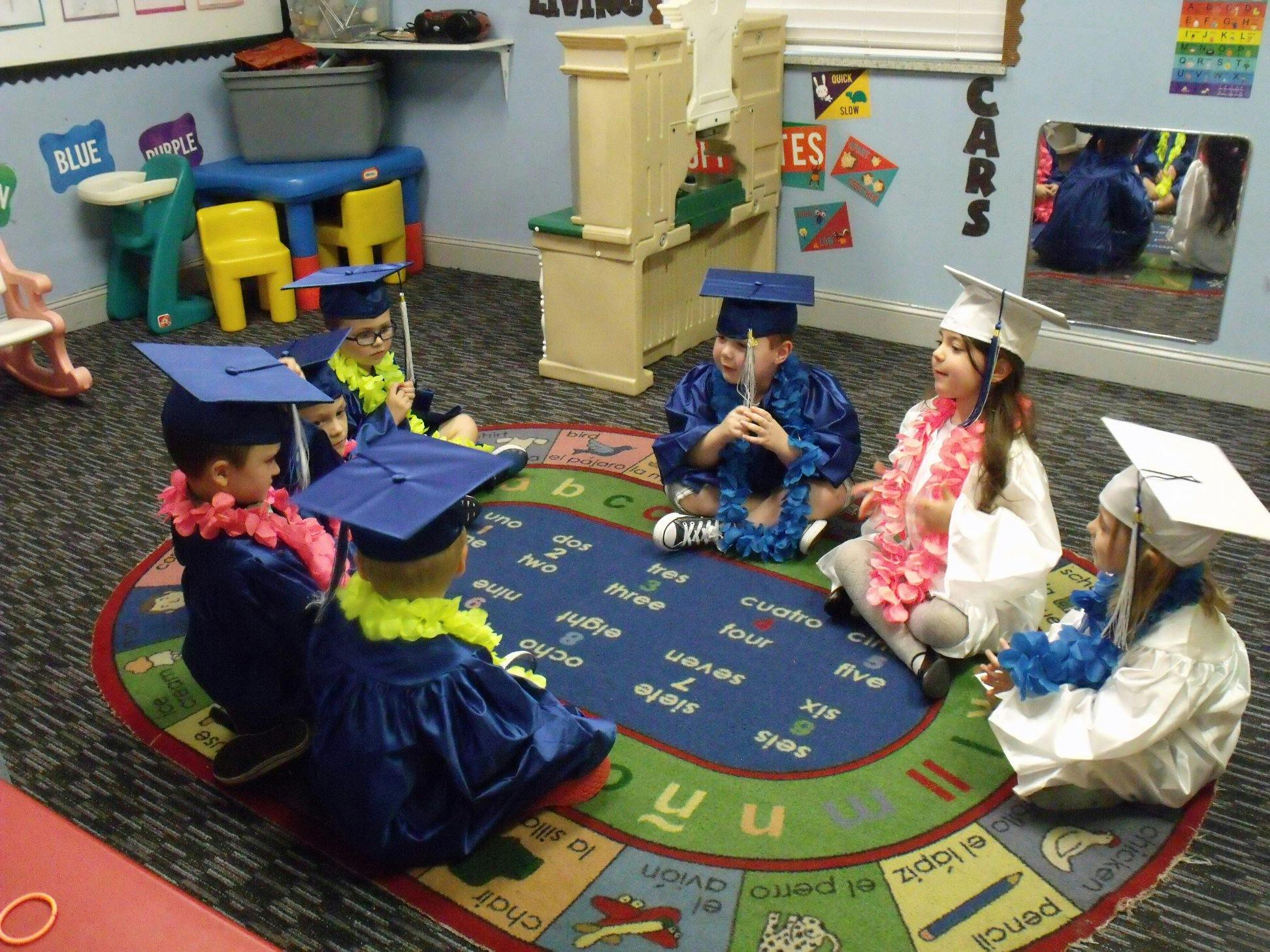 Children in graduation caps and gowns sitting on a rug in a classroom