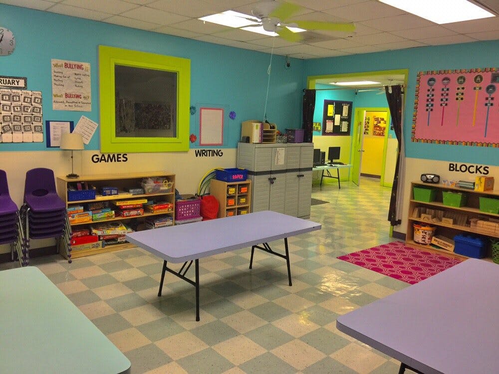 Classroom with light blue walls, tables, chairs, and shelves with supplies