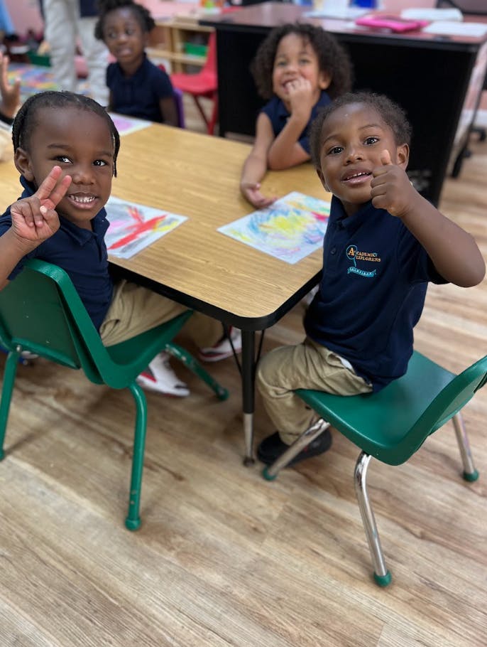 Children sitting at a table with drawings, one making a peace sign, another a thumbs up
