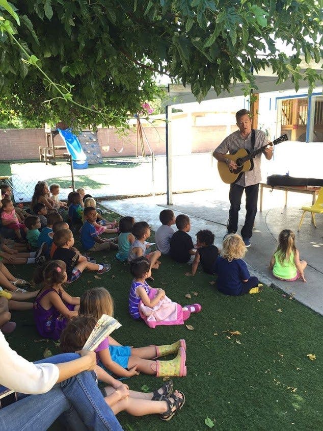 Person playing guitar for a group of children sitting on grass