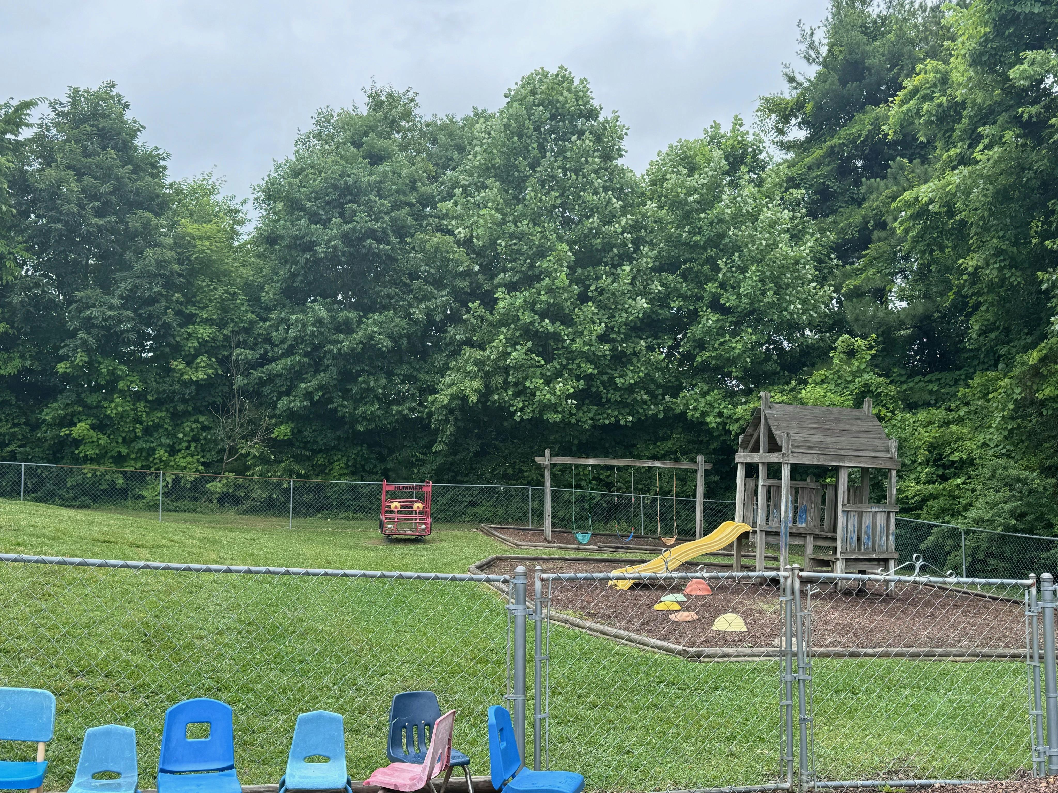 A fenced-in playground with a wooden playset, yellow slide, swings, and chairs