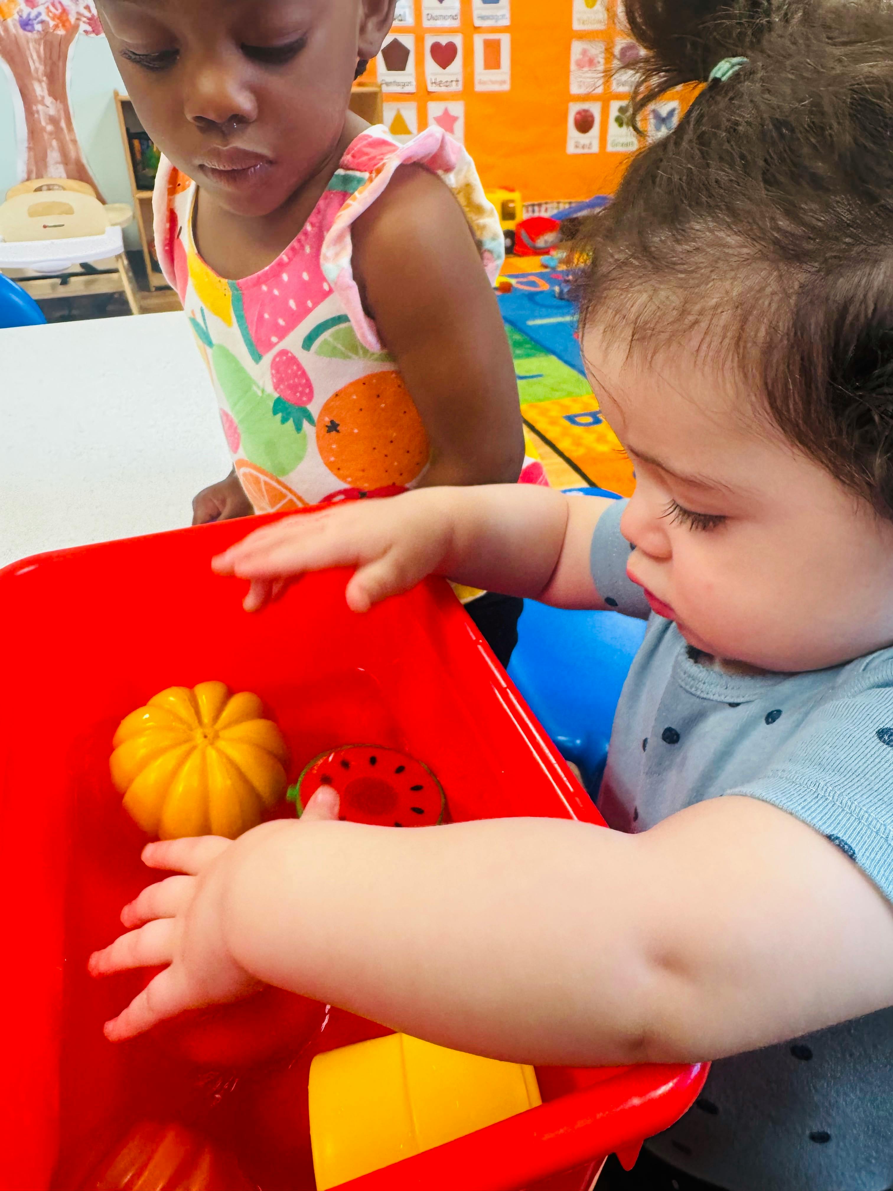 Two children playing with plastic food toys in a red water bin