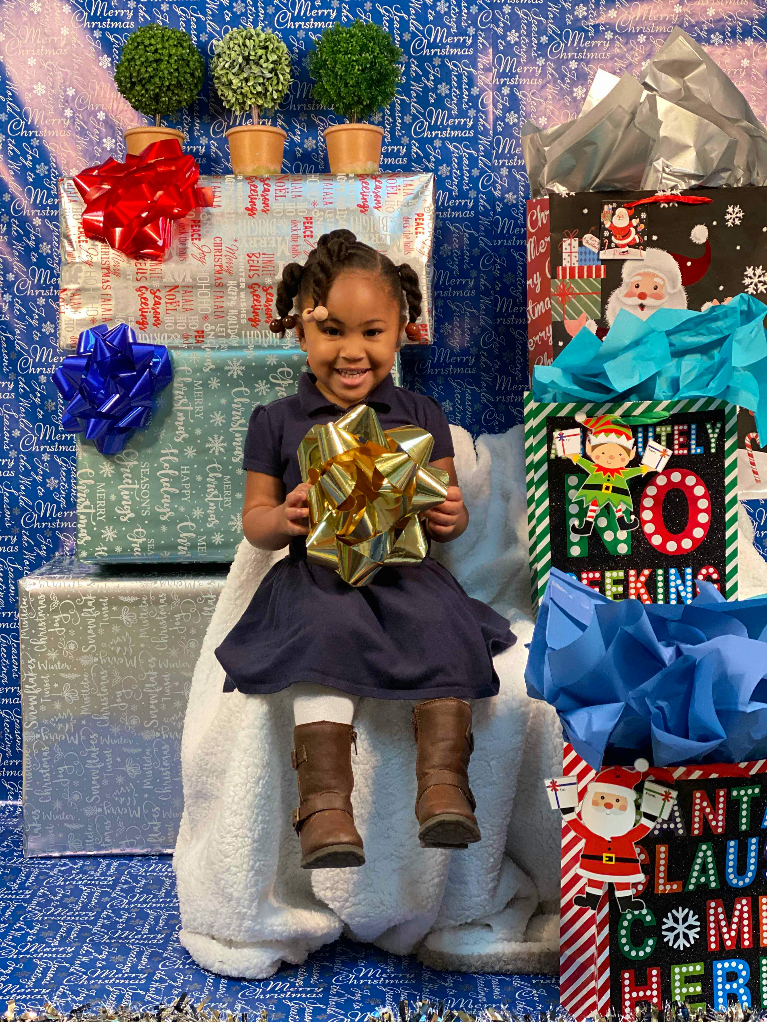 Child sitting on a white chair holding a gold bow, surrounded by wrapped gifts