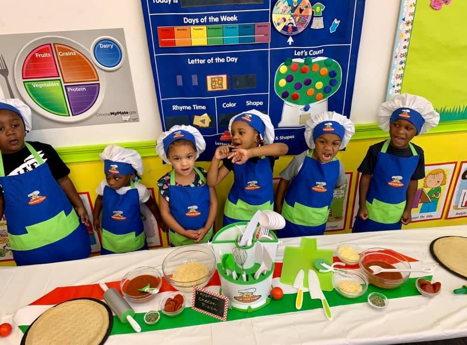 Children in chef hats and aprons standing behind a table with pizza-making ingredients