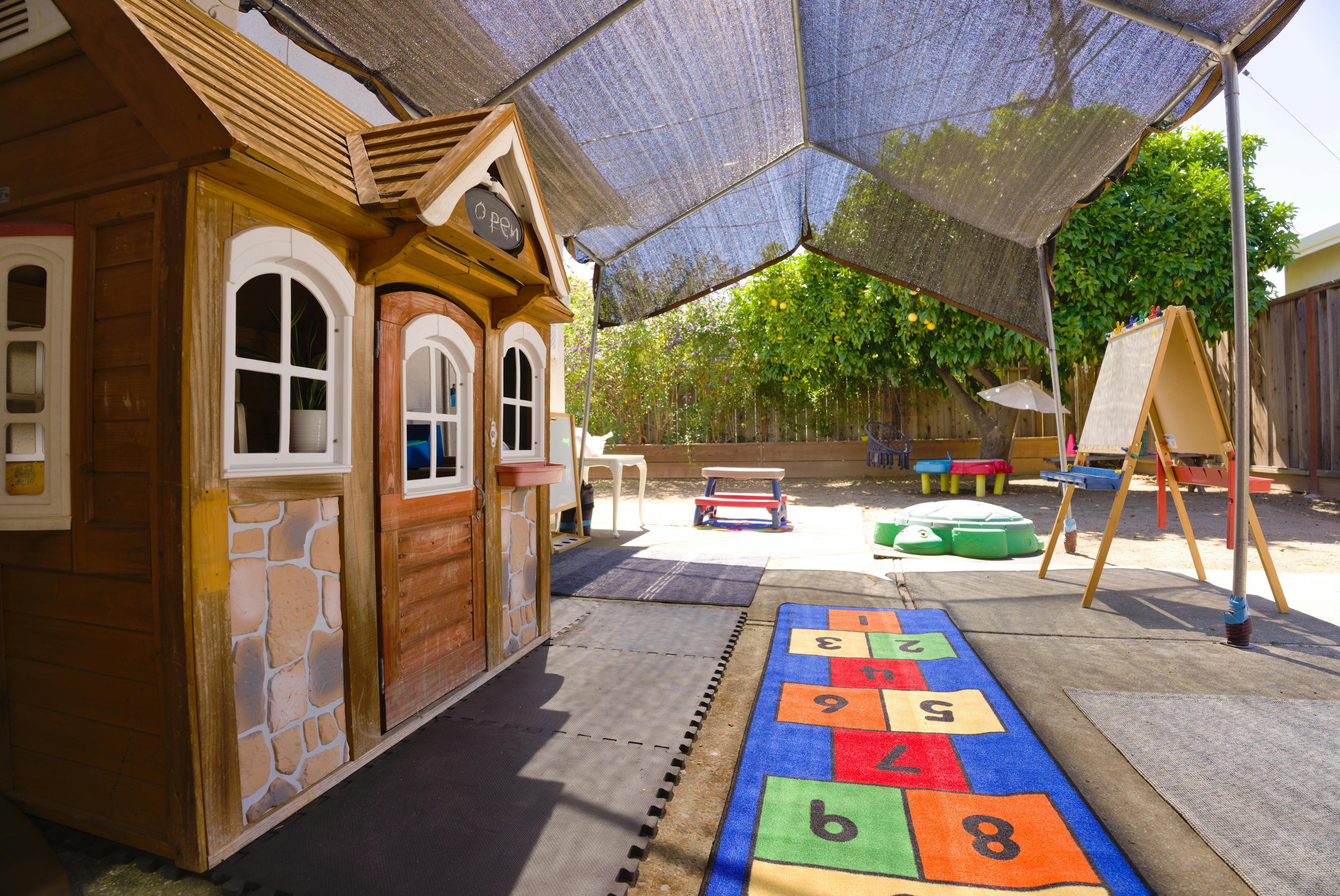 Outdoor play area with a wooden playhouse, an easel, and a hopscotch mat