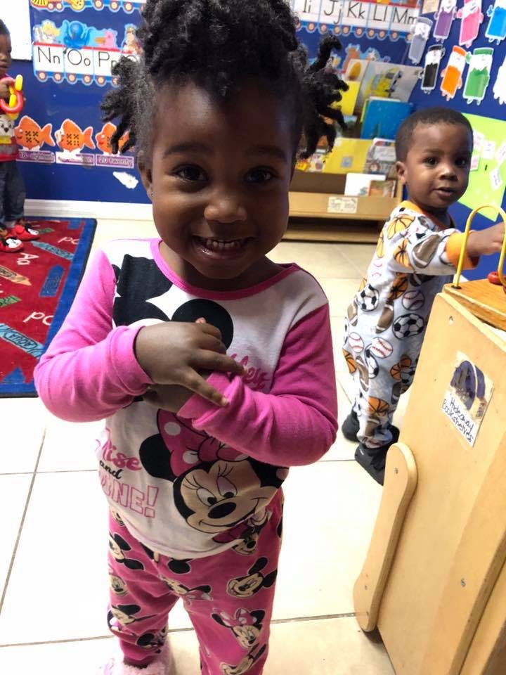 Child in pink pajamas smiling at camera, another child playing with a toy in a classroom