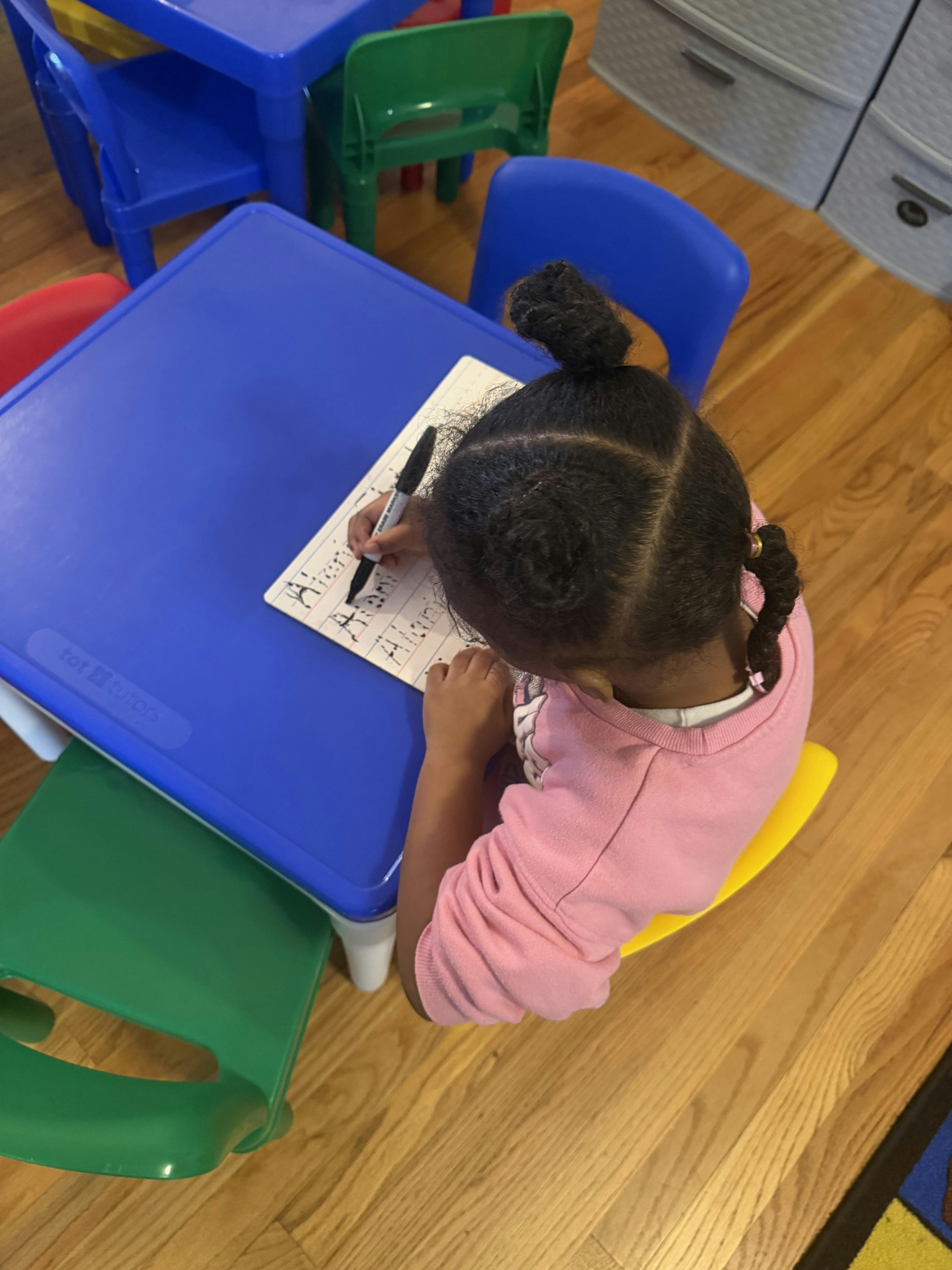child sitting at a blue table, writing with a marker on a white board