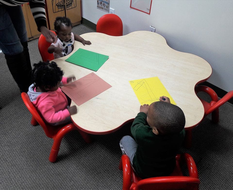 Three children sitting at a table with paper, one person standing nearby