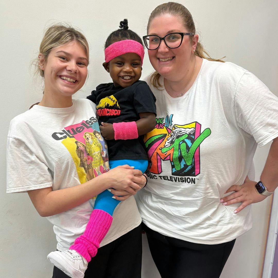 Two people holding a child dressed in 80s-style clothing