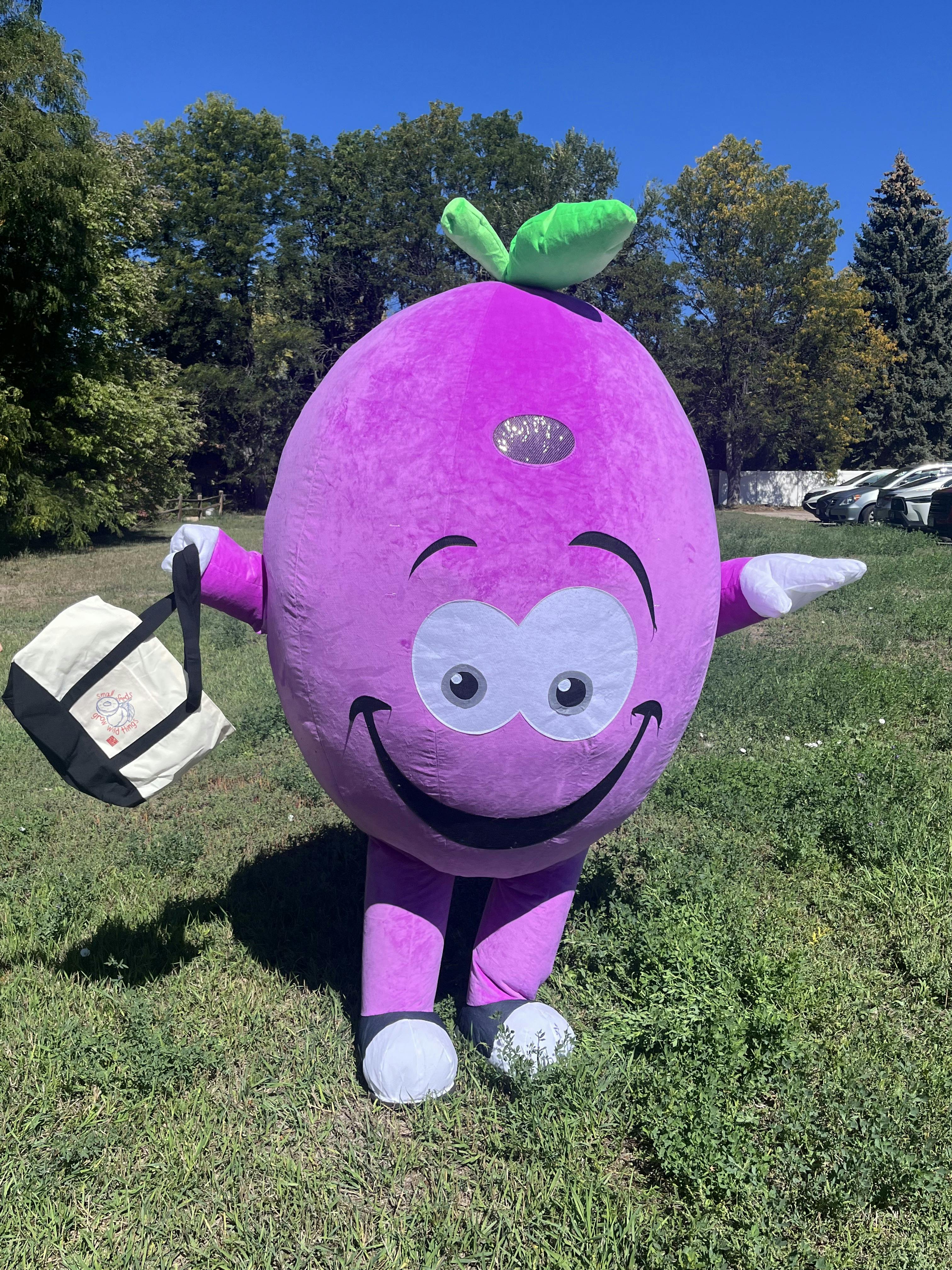 Purple mascot with a green stem, holding a tote bag
