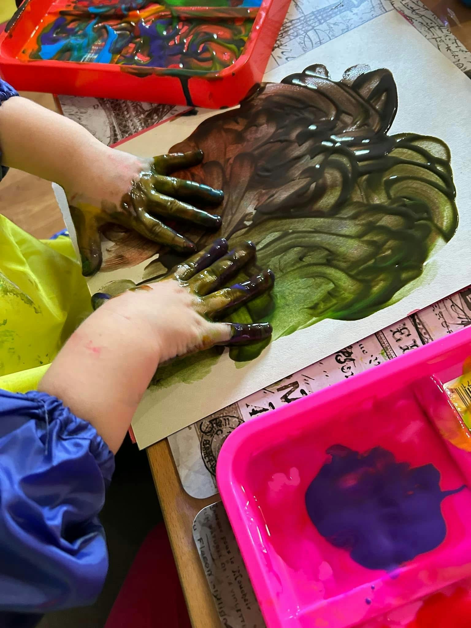 Child's hands covered in green paint on a canvas with a red paint tray