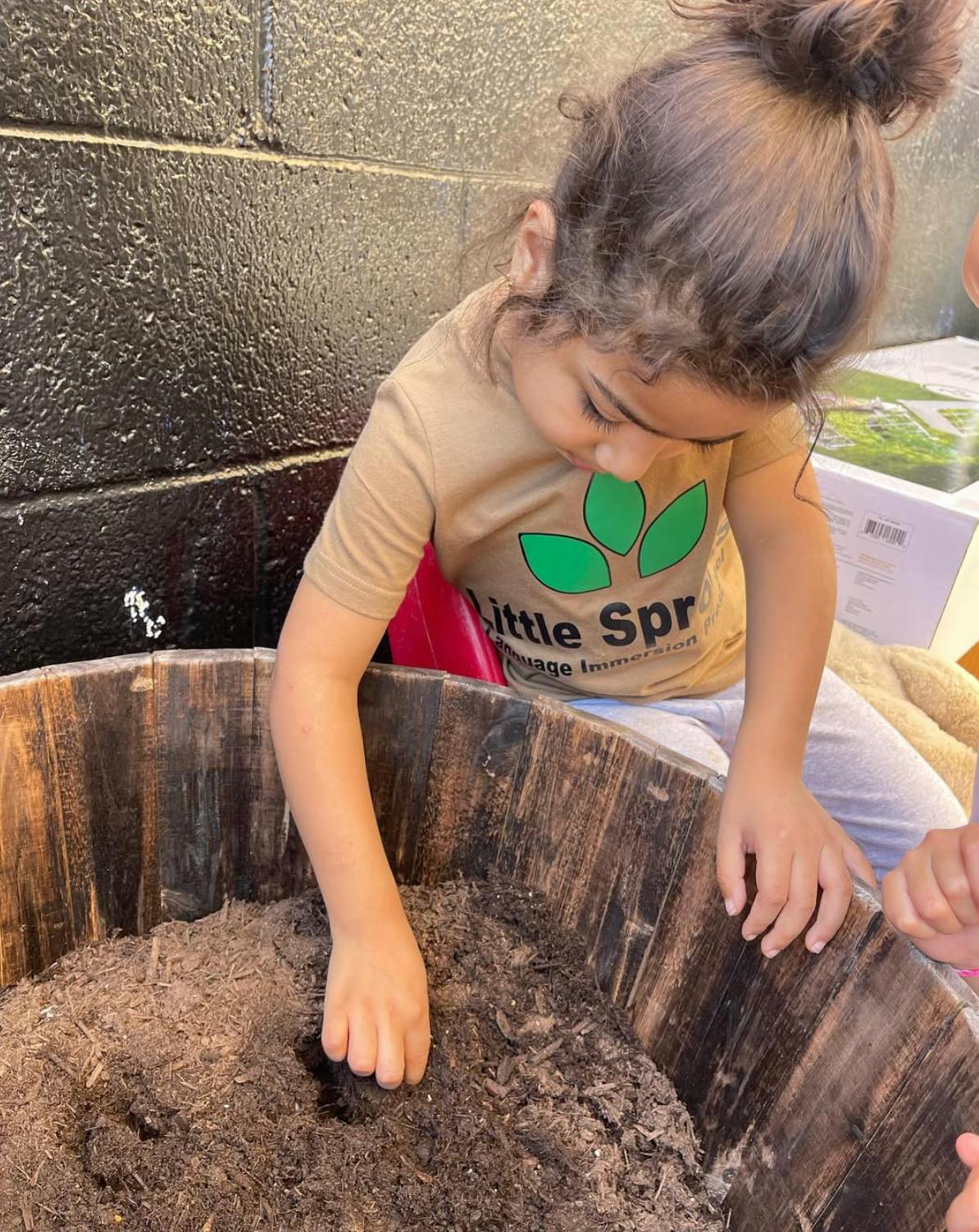 Child with hair in a bun playing in a large wooden tub of dirt