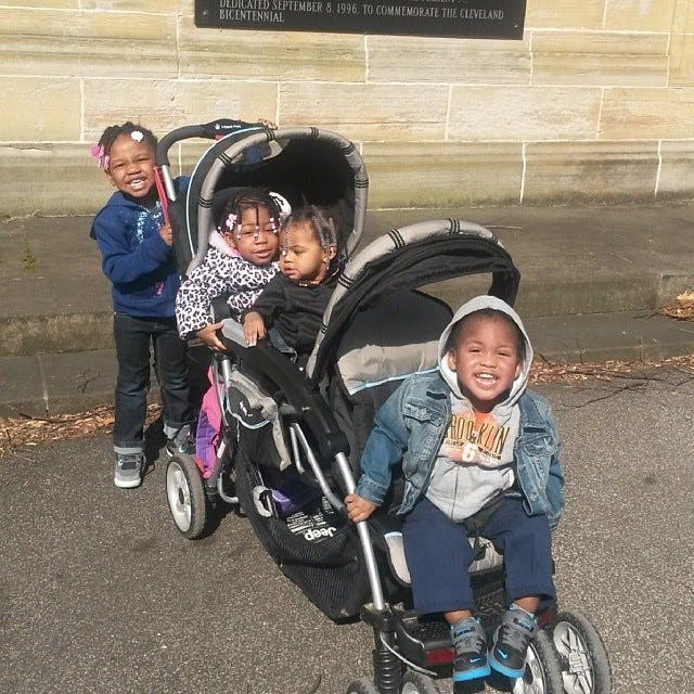 Four children with a double stroller, one child standing, three in strollers