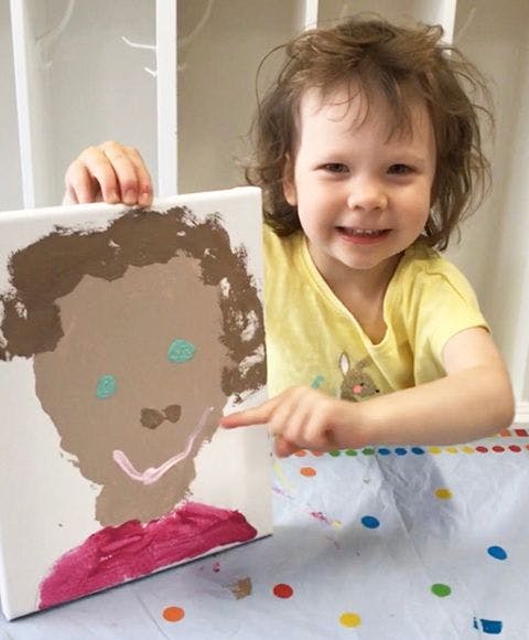 Child holding up a painting of a face and pointing to it