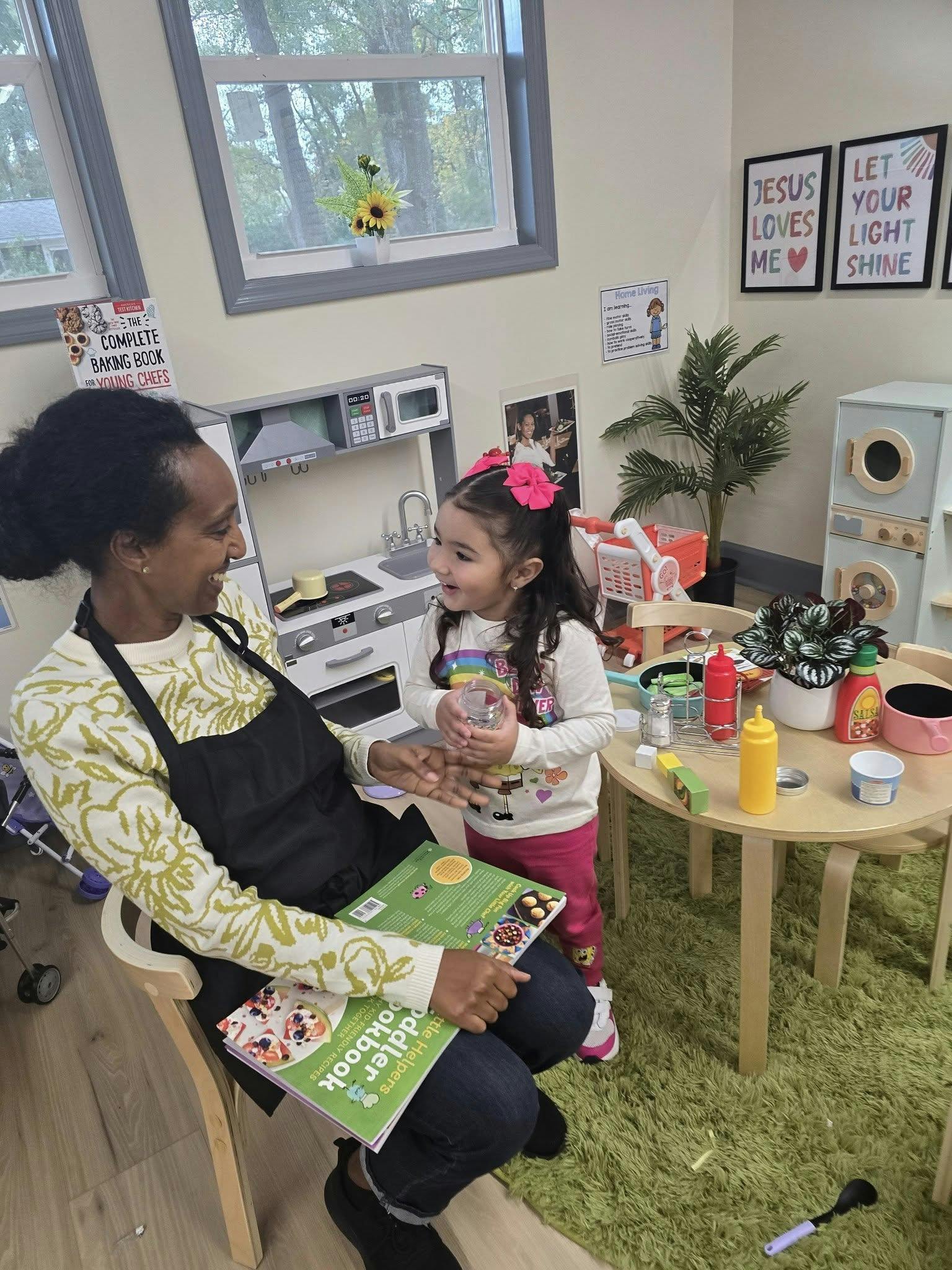 Adult and child in a play kitchen area, adult holding a "Toddler Cookbook"
