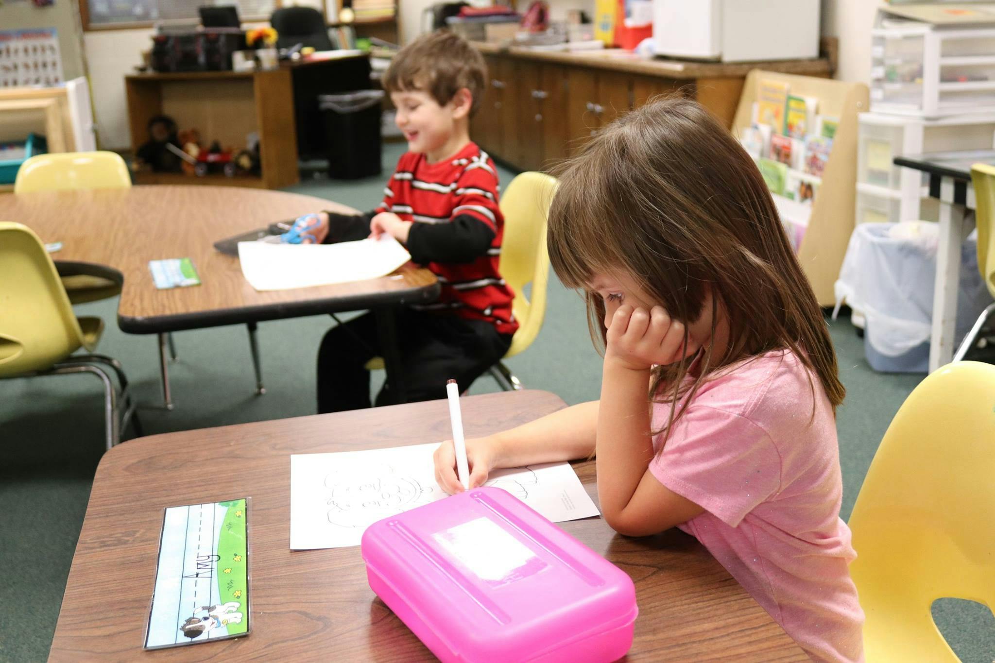 Child writing at a desk in a classroom, another child in the background