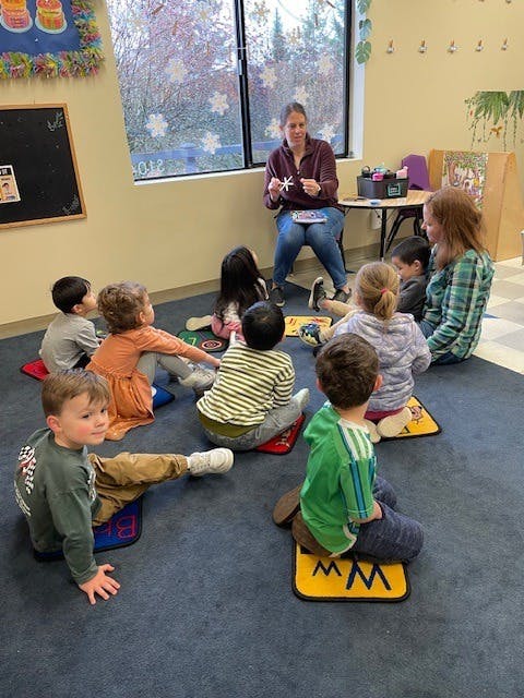 Teacher holding up a craft item for children sitting on floor cushions in a classroom