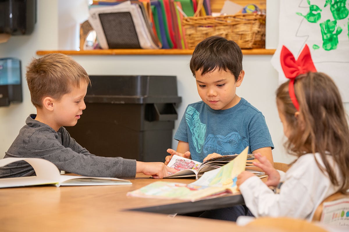Three children reading books at a table in a classroom