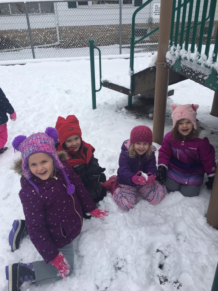 Four children in winter clothing kneeling in snow next to playground equipment