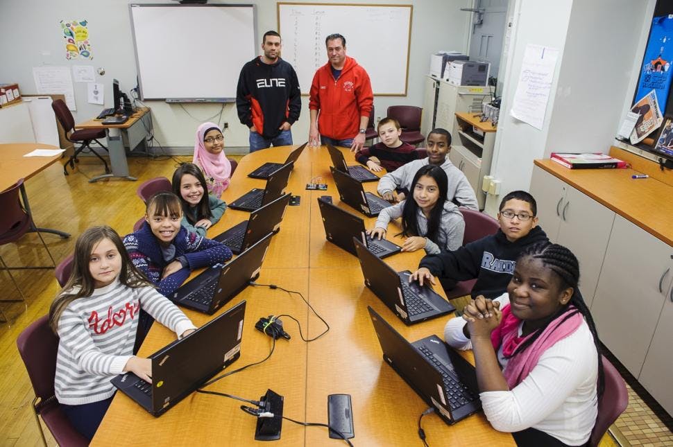 Two adults and multiple children in a classroom with laptops
