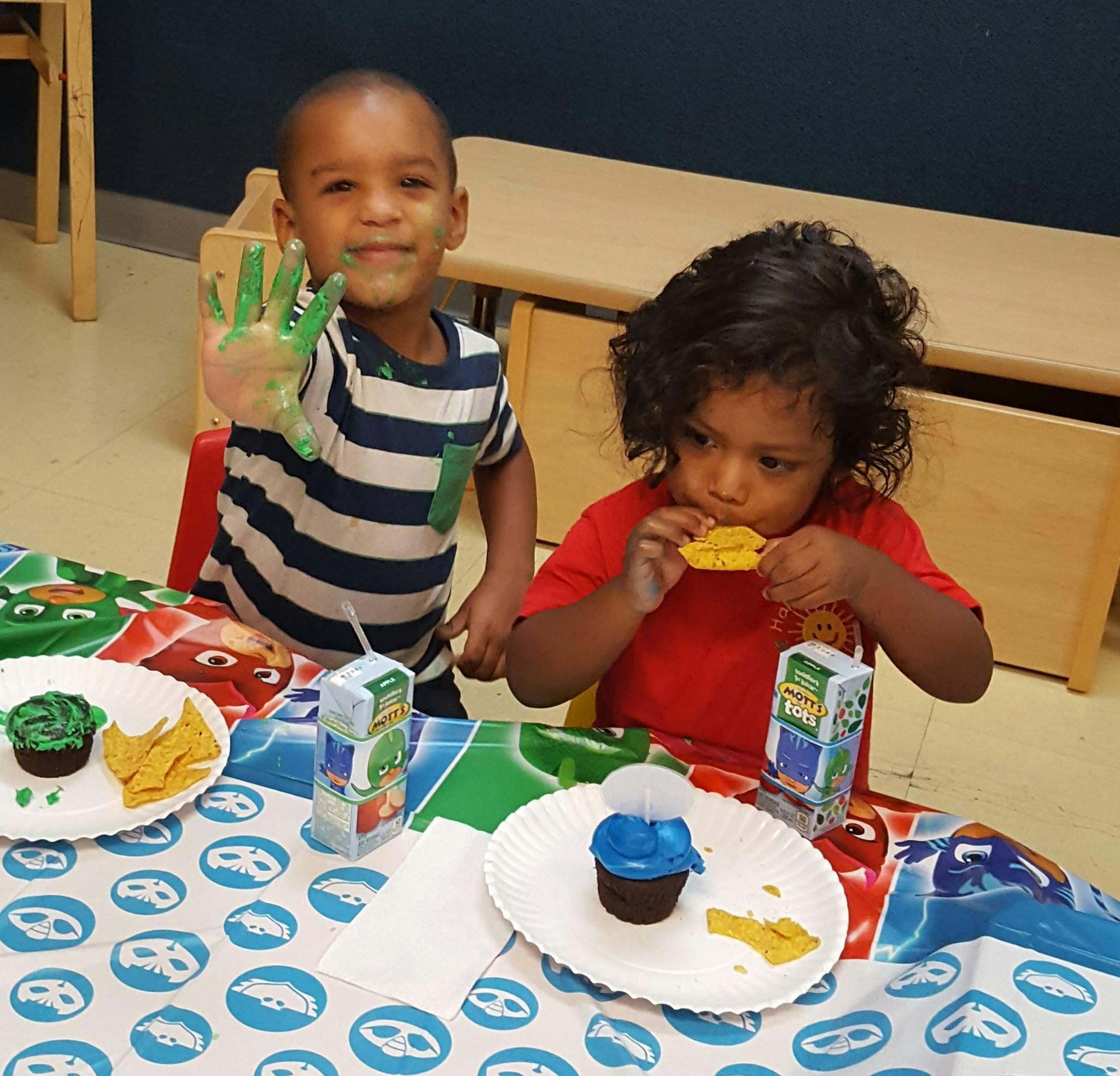 Two children at a table, one with green paint on hand, one eating