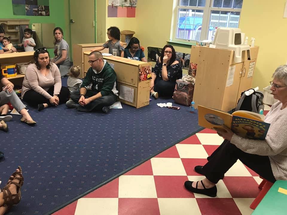 Adults and children sitting on the floor listening to a person read a book
