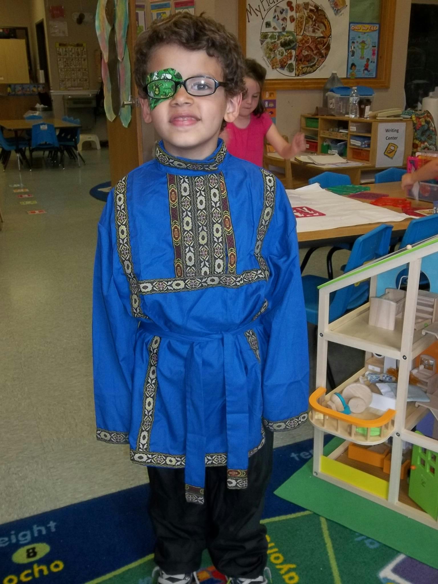 Child wearing glasses and an eye patch, dressed in a blue tunic
