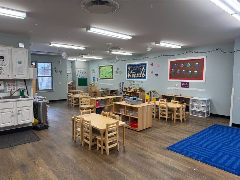 Classroom with small wooden tables and chairs, shelving units with toys, and wall decorations