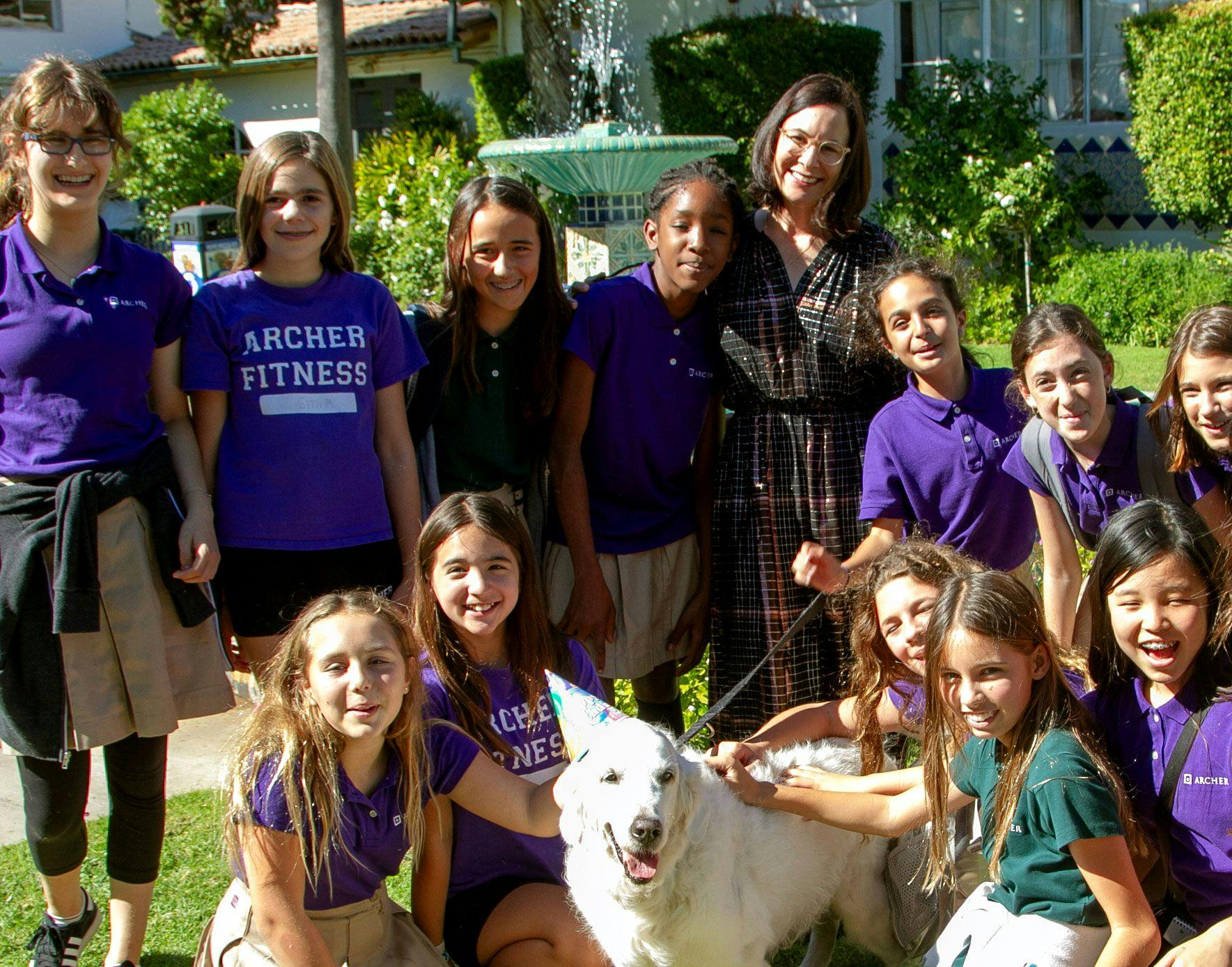 Group of children and one adult standing with a white dog wearing a party hat