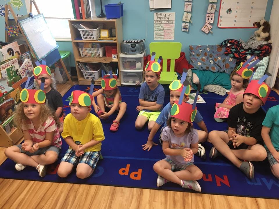Children wearing colorful hats sitting on a blue rug in a classroom