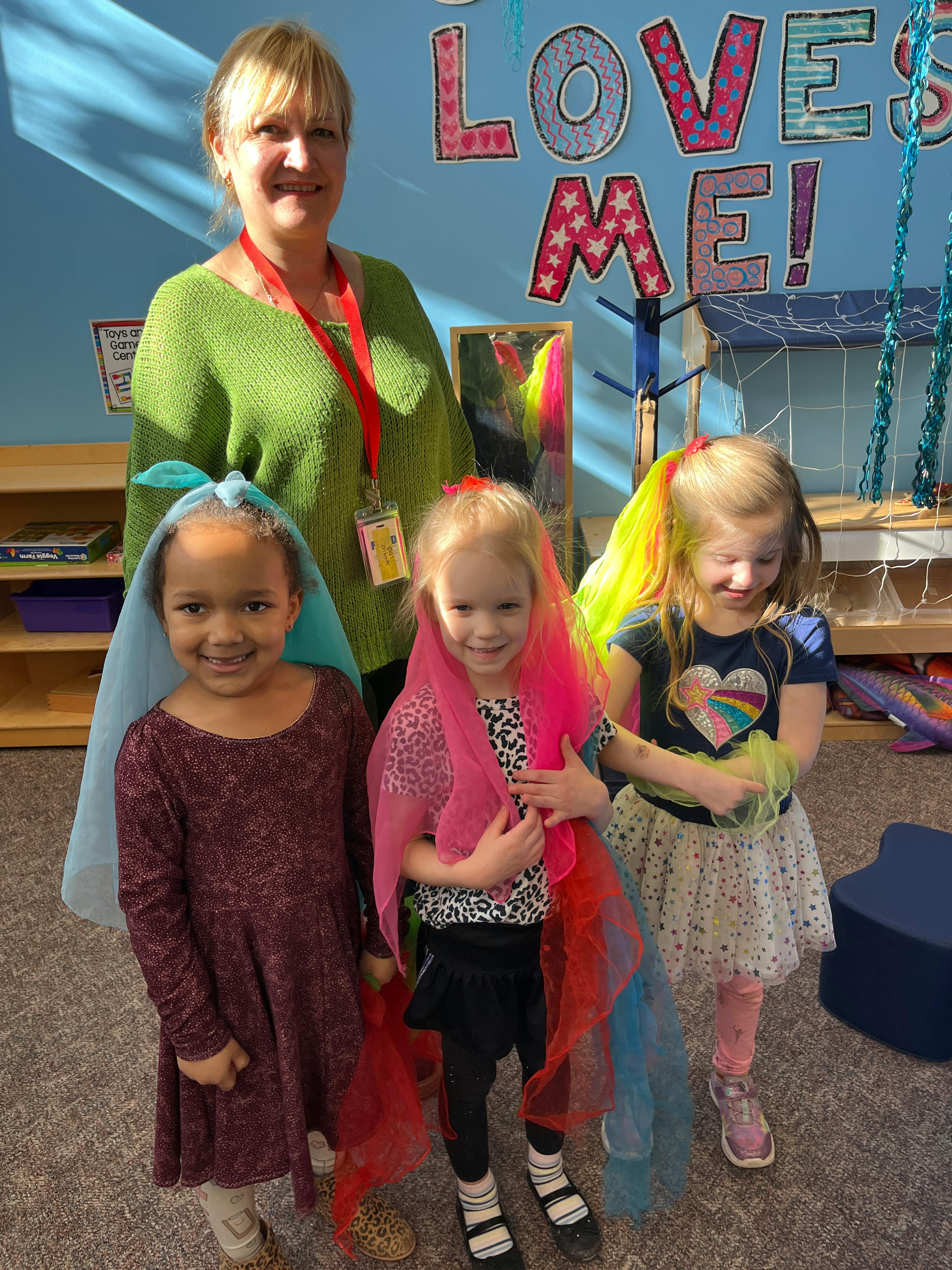 Adult and three children in a classroom, two children wearing colorful scarves